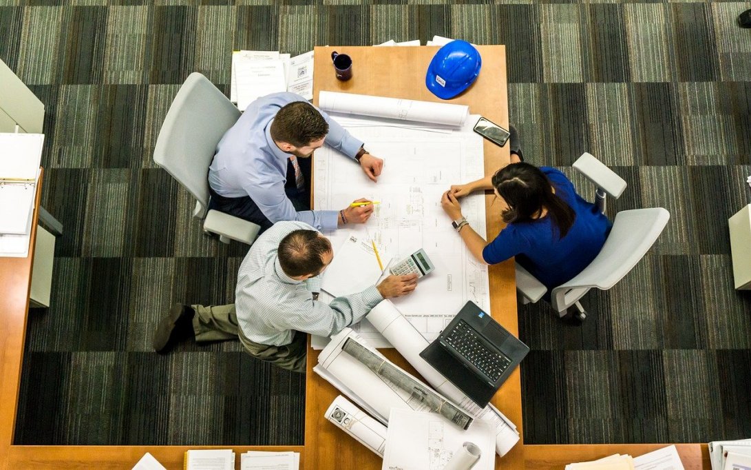 Overhead view of three people sitting at a conference table working together. The workers have papers spread out, there is also a hard hat, laptop, phone, and calculator on the table among them.